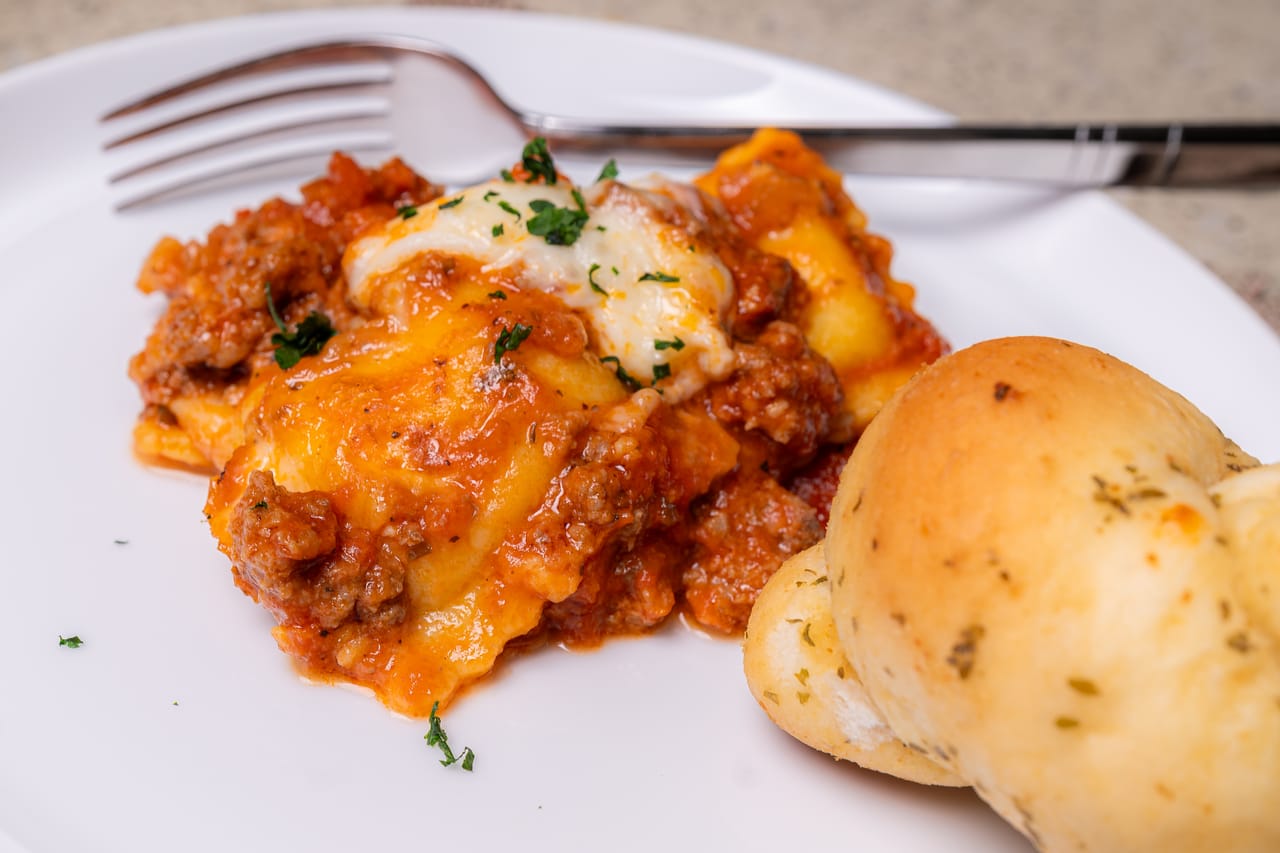 Baked ravioli in meat sauce with melted cheese and parsley on a white plate, with a garlic knot on the side