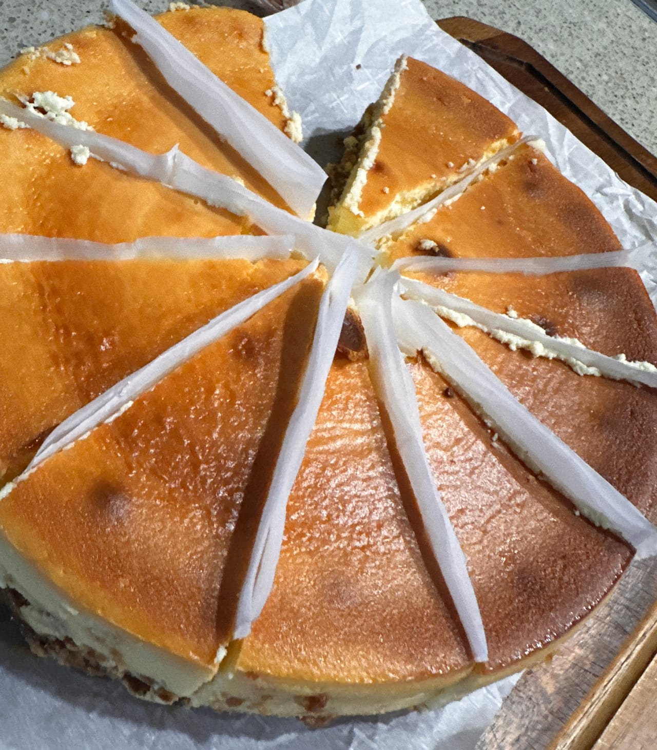 A golden-topped baked cheesecake cut into wedges, with strips of paper between the slices, on a wooden board