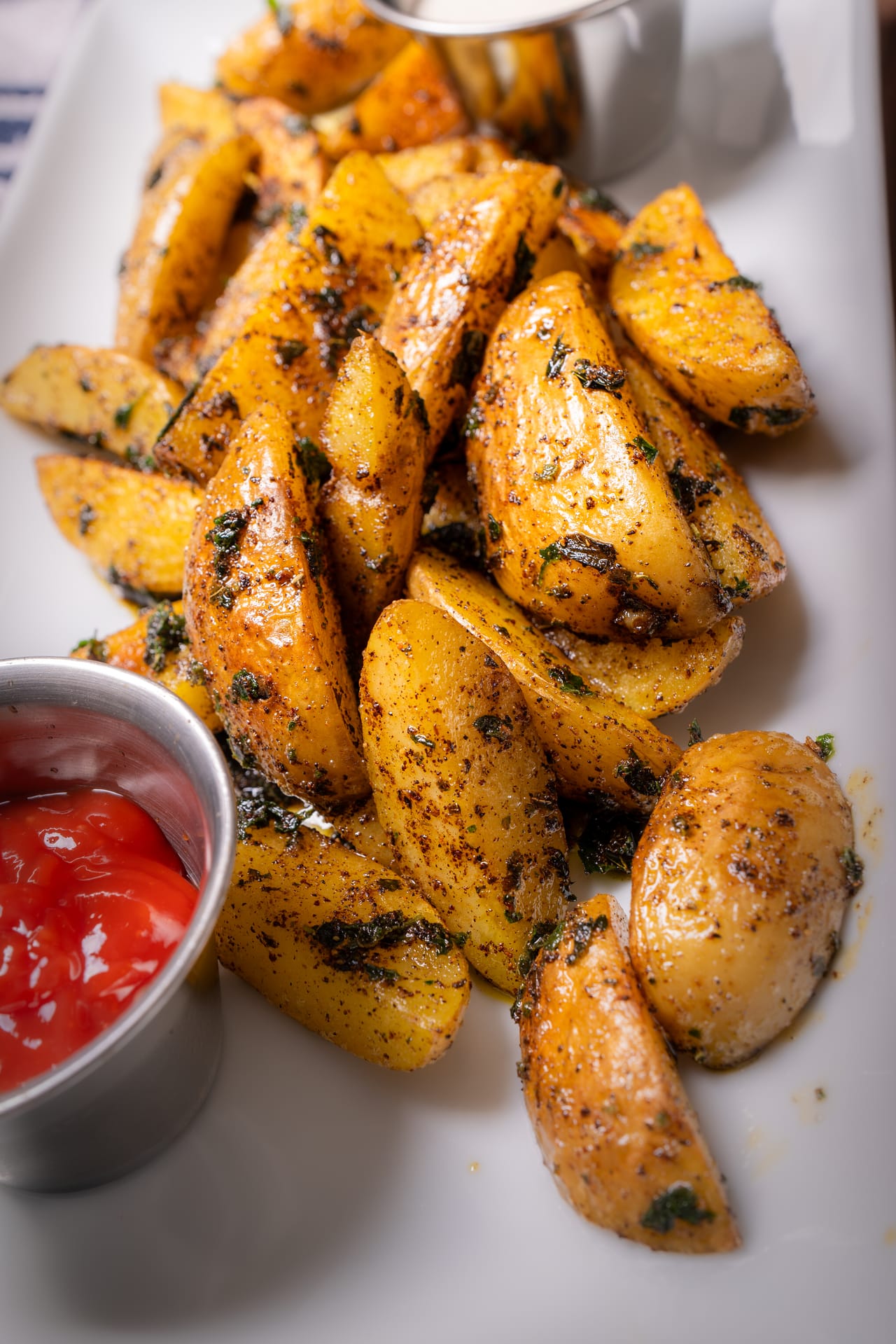 Golden potato wedges with herbs on a white platter, next to a cup of ketchup and a white dip
